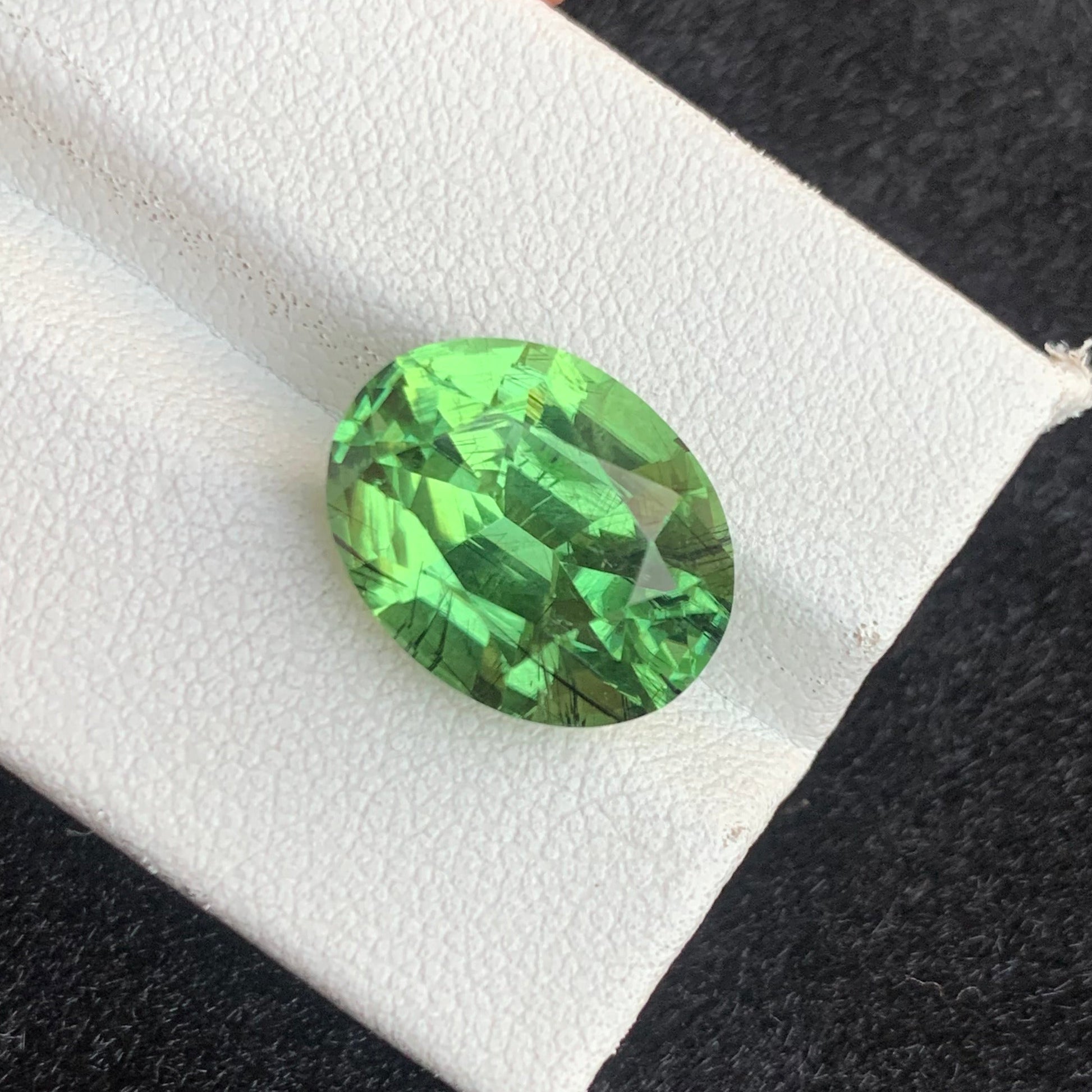 A small oval-cut bright green gemstone displayed on a white textured card held by fingers against a dark black background.